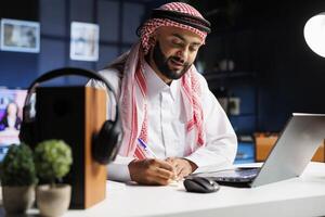 An engrossed male entrepreneur, dressed professionally, sits at a desk with his personal computer. Muslim man surfs the net on his laptop, surrounded by a notepad and focused on his online research. photo