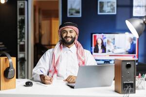 Middle Eastern male freelancer in traditional attire, browsing the internet on laptop while holding a pen. Arab man smiles confidently at camera, reflecting his professional profile in modern office. photo