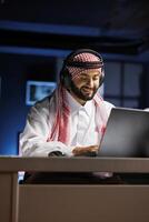 Arab guy typing confidently while using a laptop for email, conferencing, and conversation. Muslim man studying and surfing the web while using earphones to listen and watch. photo