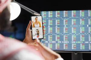 Muslim man holding his smartphone and participating in online meeting, symbolizing integration of technology in the workplace. Over-the-shoulder shot of mobile device held in front of computer screen. photo
