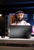 Middle Eastern man in traditional attire efficiently uses a laptop for research and communication via email. Arab guy with wireless headphones having a call on his personal computer. photo