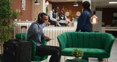 Client listens to music at hotel reception, preparing to sign registration papers waiting to do check in at front desk in lobby. African american person using headset in lounge area, tourism. photo