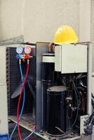 Close up of damaged external opened out of service air conditioner unit in need of fixing. Yellow protection helmet on top of outdoor HVAC system with manifold gauges, compressors, wiring photo