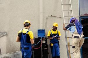 Diverse team of knowleadgeable engineers using pressure measurement device to check liquids and gases in air conditioner and write inspection and safety check report on clipboard photo