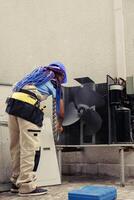 African american skilled mechanic commissioned by home owner for annual air conditioner routine maintenance, using soft dusting brush to clean dusty condenser coils and filters, outdoor photo