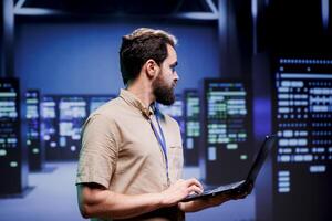 IT developer in server hub storing data management rigs, storage devices and networking devices offering vast amounts of computing resources. Employee doing infrastructure maintenance using laptop photo