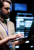 Serviceman doing system analysis, typing data on laptop of needed adjustments in supercomputers server farm housing advanced data storage infrastructure supporting critical IT workloads, close up photo