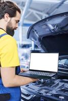 Qualified technician in garage using mockup laptop to follow checklist while doing maintenance on car. Skilled worker in auto repair shop does checkup on vehicle helped by isolated screen device photo