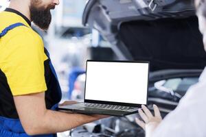 Certified engineer in garage using mockup laptop to follow checklist while doing maintenance on car. Adept worker in auto repair shop does checkup on vehicle helped by isolated screen device photo