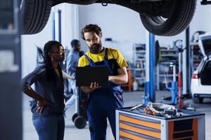 Trained mechanic in garage using laptop to follow checklist while doing maintenance on car, talking with client. Specialist in auto repair shop does checkup on vehicle motor next to customer photo