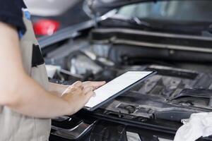 Certified mechanic in garage using tablet to follow checklist while doing maintenance on car. Close up shot of specialist in auto repair shop doing checkup on vehicle engine photo
