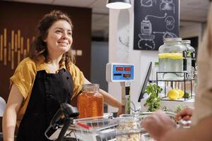 Cheerful woman at checkout counter in zero waste shop selling healthy lentils in jar to customer with green living lifestyle. Client buys food in local neighborhood store from smiling vendor photo