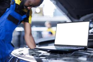 Mockup laptop on car with hood open while serviceman in blurry background replaces radiator. Isolated screen device next to knowledgeable garage worker fixing client vehicle photo