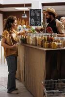 Client shopping for organic pantry staples in zero waste store using biodegradable paper bag, being assisted with information by trader. Vendor showing woman products in reusable jars photo