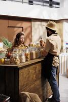 Client shopping for organic pantry staples in zero waste store using plastic free paper bag, being assisted with information by trader. Seller showing woman products with no artificial ingredients photo