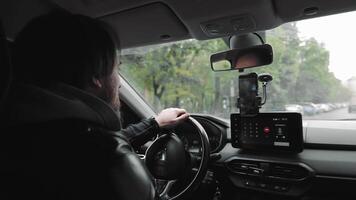 Man driving car while using hands-free phone. A man in a leather jacket drives a car while talking on a hands-free phone mounted on the dashboard on a tree-lined street. video