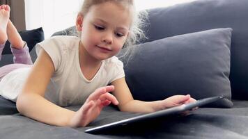 Little girl using tablet device, lying on sofa. Smiling little girl watching online cartoons on tablet on Internet using digital tablet technology device. . High quality 4k footage video