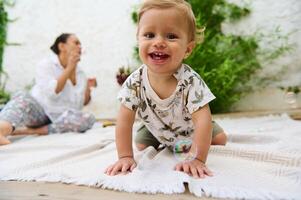 Portrait of a cute little baby boy smiling at camera while crawling on a linen blanket in the backyard. Happy mother blowing soap bubbles on blurred background. Carefree babyhood and infancy concept photo