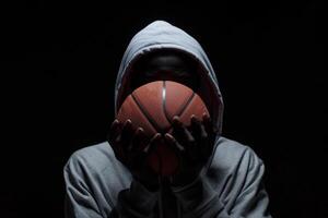 Mysterious figure in hoodie holding basketball, shadows and light playing across face emphasizing the sport's intensity and style. photo