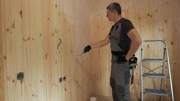 This shows a worker applying acrylic paint to a wooden wall in a new house. The worker is wearing work clothes and gloves. There is a ladder in the background. video