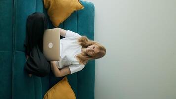 Happy 30s woman making call on laptop while relaxing on a sofa having pleasant conversation waving hand greeting gesture smiling to laptop screen video