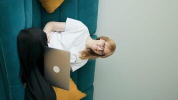 Happy 30s woman making call on laptop while relaxing on a sofa having pleasant conversation waving hand greeting gesture smiling to laptop screen video
