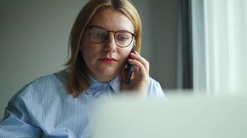 Business young female employee speaking on telephone and using app laptop at office workplace. Multitasking manager talking on mobile phone, making call to client, advisor giving consultation video