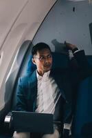 Smart and focused Asian businessman in formal business suit using laptop computer during the flight for his business trip. Business people and transportation concept photo
