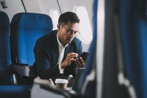 Attractive Asian male passenger of airplane sitting in comfortable seat while working laptop and tablet with mock up area using wireless connection. Travel in style, work with grace. photo