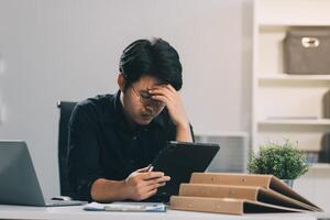 Frustrated Asian young businessman worker working on table in office. Busy employee worker people feel tired and headache from late work and overwork job document on computer desk at company workplace photo