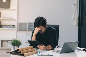 Frustrated Asian young businessman worker working on table in office. Busy employee worker people feel tired and headache from late work and overwork job document on computer desk at company workplace photo
