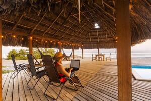 Man with a hat sitting on a chair and working on a laptop in front of the beach. photo