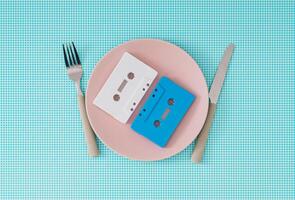 Audio cassette tapes on pink plate with knife and fork on white and blue background. Minimal food with retro music idea. Creative concept of retro technology. 80's or 90's aesthetic vintage style. photo