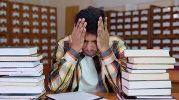 Tired indian student, in casual clothes, sits at a desk in a library between books, holds his hands on his head, looks tired of reading video