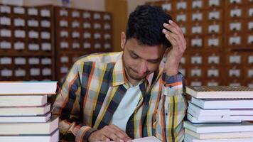 Tired indian student, in casual clothes, sits at a desk in a library between books, holds his hands on his head, looks tired of reading video