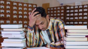 Tired indian student, in casual clothes, sits at a desk in a library between books, holds his hands on his head, looks tired of reading video