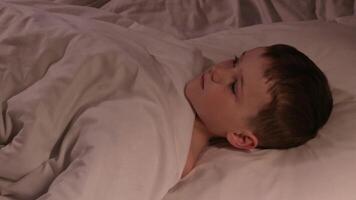 kid boy with eyes open cannot sleep at night in a bed in the bedroom and dreams. Child son looks at ceiling on a pillow under a blanket in evening video