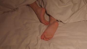 restless feet of a kid boy on a sheet under a blanket at night. Baby child girl with bare legs tosses and does not fall asleep in bed in the bedroom in evening video