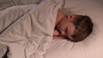 Cute child boy wakes up after sleeping in the morning in bed in the bedroom. Happy child son is lying on a pillow under blanket and smiling video