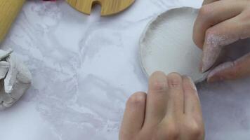 Person shapes circle of clay by bending edges. Piece of white clay for modeling on marble table. Preparation of material for manufacture of ceramic products. Handmade. Wooden tools on background. video