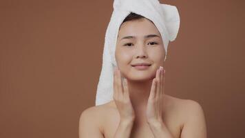 jung attraktiv asiatisch Frau mit sauber Haut und ein Handtuch auf ihr Kopf bewirbt sich ein feuchtigkeitsspendend Sahne auf ihr Wangen nach ein Dusche und lächelnd. Konzept von Haut Pflege zum Gesundheit und Schönheit. video