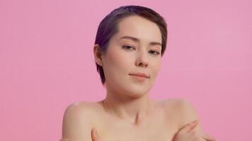 Young attractive woman with clean skin touching her shoulders with her hands close up and coquettishly looking at the camera on a pink background. Concept of skin care for health and beauty video