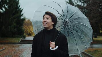 Front view of a young man in a coat walking under an umbrella during a rain in the autumn city park, looking at the sides and smiling. Concept of comfort leisure in bad weather video