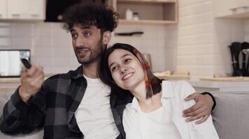 Young man in casual clothes sitting on a couch and hugging a smiling woman while watching TV in a cozy kitchen at home. Concept of happiness and harmony in a couple relationship video