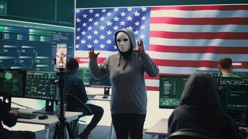 Person in anonymous mask working on intelligence in a governmental hacking room, recording himself and asking for ransom with the USA flag on big screen. Cyber defense information security. Camera B. video