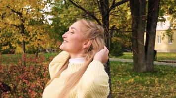 Close-up of young blonde Caucasian woman standing in park, looking upward smiling with both hands behind head with trees covered with sun in background. Autumn walk in park. video