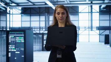 Software developer walking in data center, overseeing server operations using laptop. Woman ensuring optimal performance and data integrity in server farm using notebook, camera A video