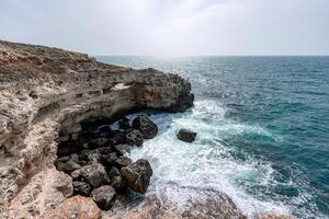 The ocean is calm and the sky is cloudy. The water is a deep blue color. The rocks are jagged and the waves are small. photo