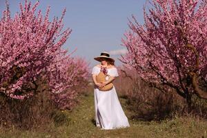 Woman blooming peach orchard. Against the backdrop of a picturesque peach orchard, a woman in a long white dress and hat enjoys a peaceful walk in the park, surrounded by the beauty of nature. photo
