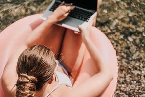 A woman is sitting in a pink inflatable pool with a laptop on her lap. She is typing on the laptop, possibly working or browsing the internet. Concept of relaxation and leisure. photo